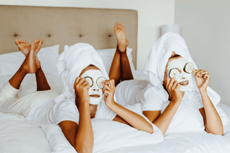 Two moms relaxing on a bed with face masks, towels on their heads, and cucumbers over their eyes during a self-care spa day.