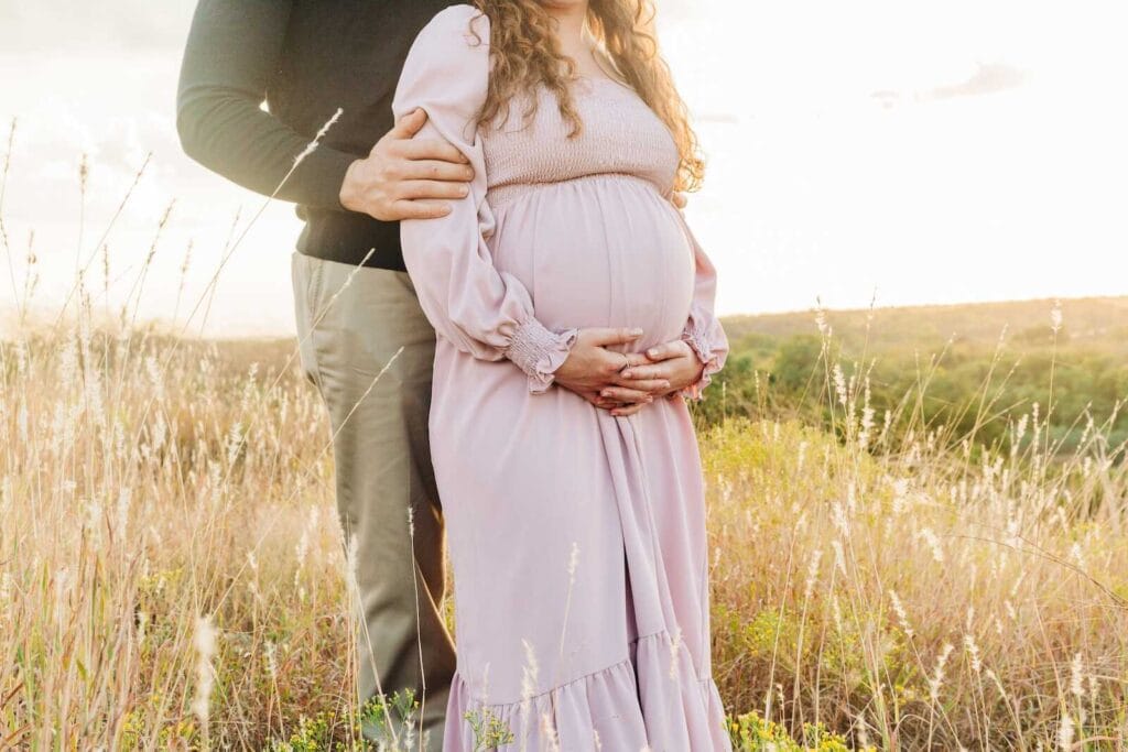 Pregnant woman holding her belly while partner stands beside her in a field.
