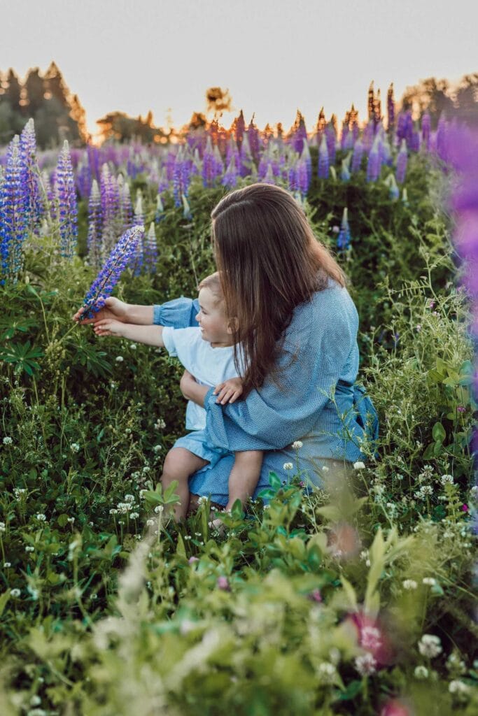 Mom sitting in a flower field holding her child and pointing at purple flowers.