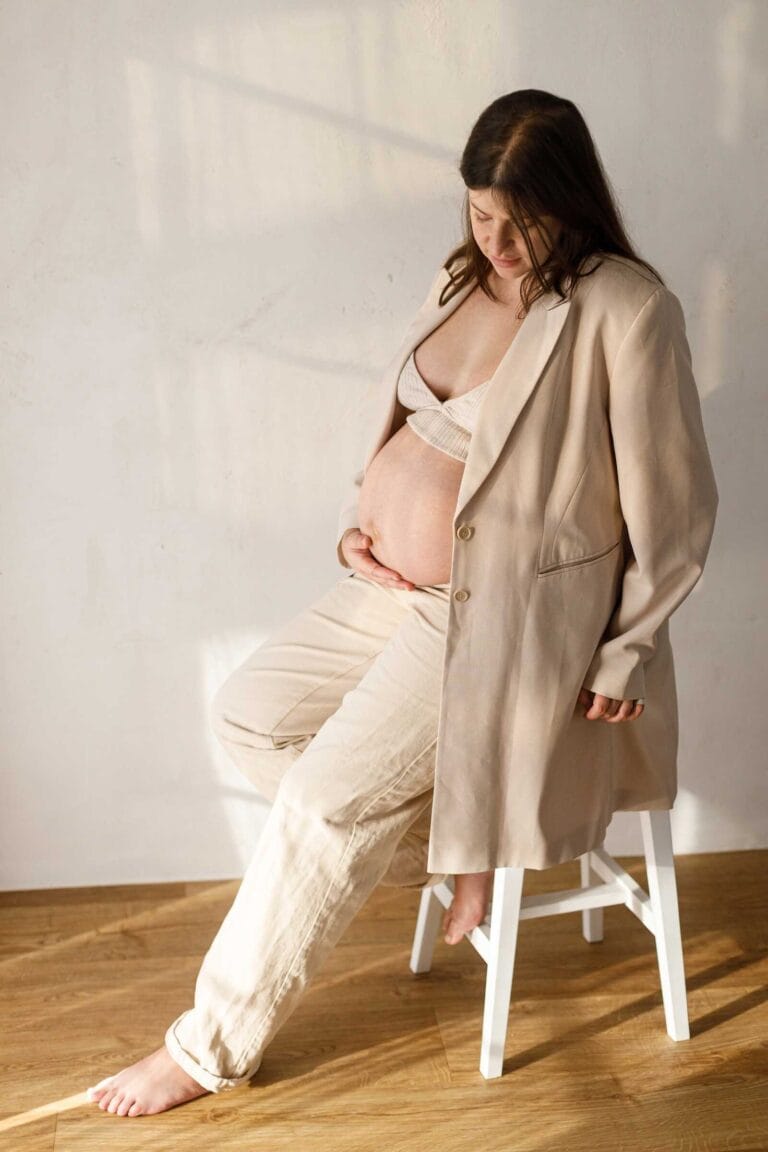 Minimalist maternity photos of pregnant woman sitting on stool in warm natural light.
