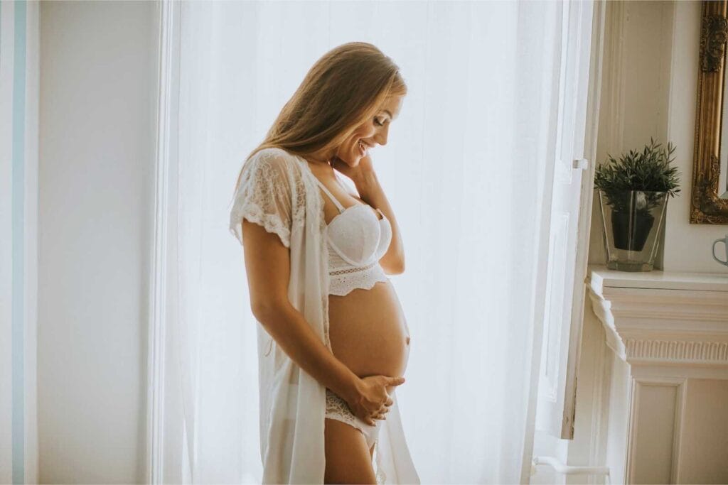 Elegant maternity boudoir photo of a pregnant woman in lace lingerie standing by a window.