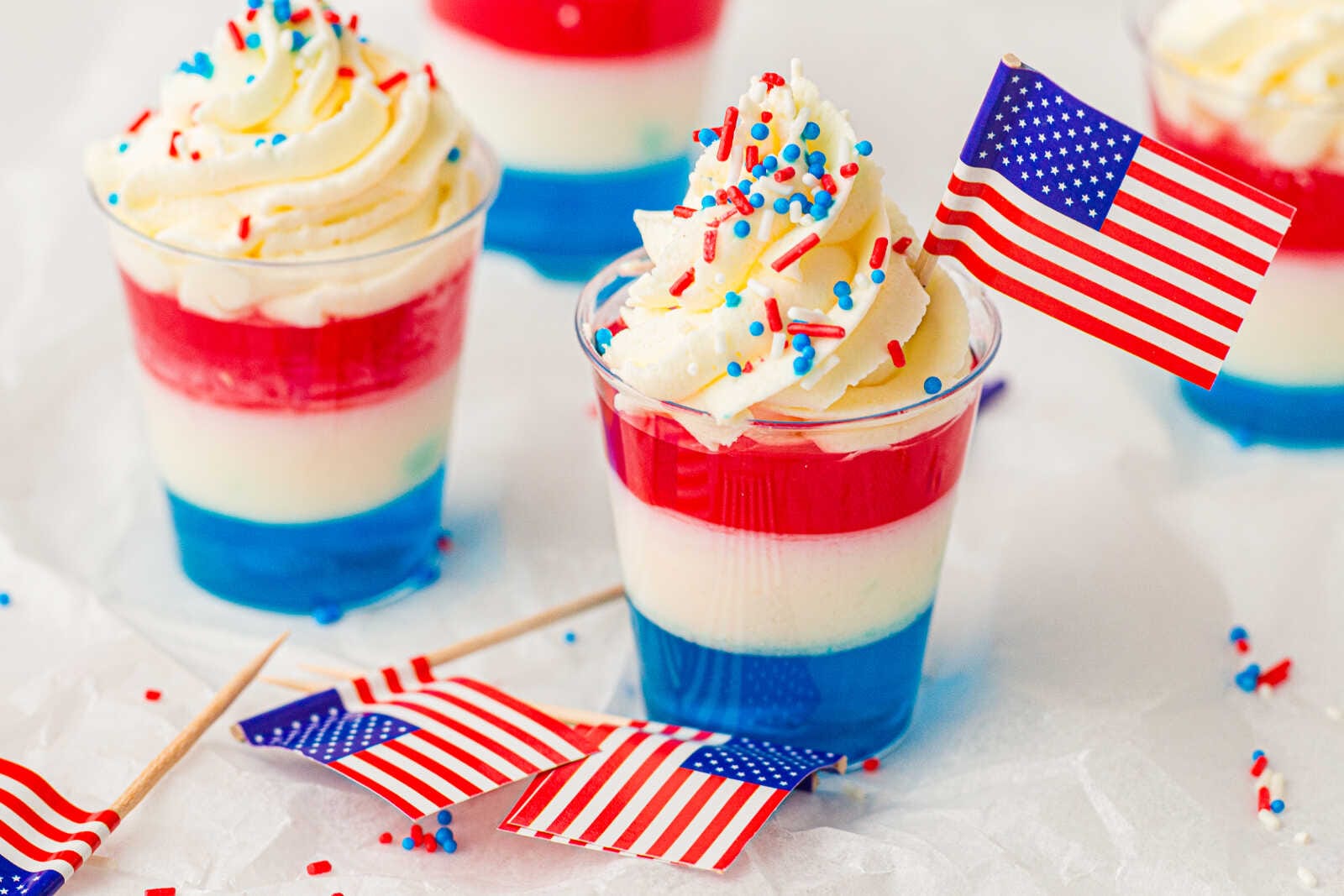 Patriotic red, white, and blue Jello cups topped with whipped cream, sprinkles, and mini American flags.